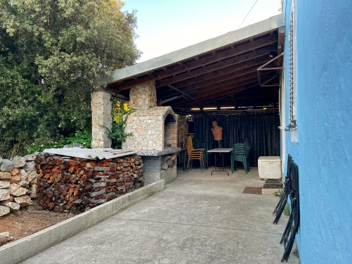 Covered terrace beside the blue house with a stone outdoor fireplace, stacked firewood, a table and chairs